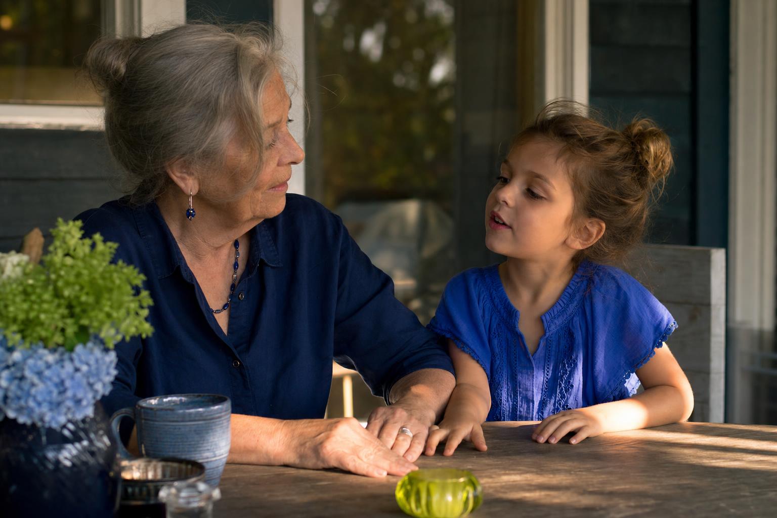 A grandmother and her young granddaughter sit together at an outdoor table in warm evening light, holding hands and talking, a vase of hydrangeas beside them.